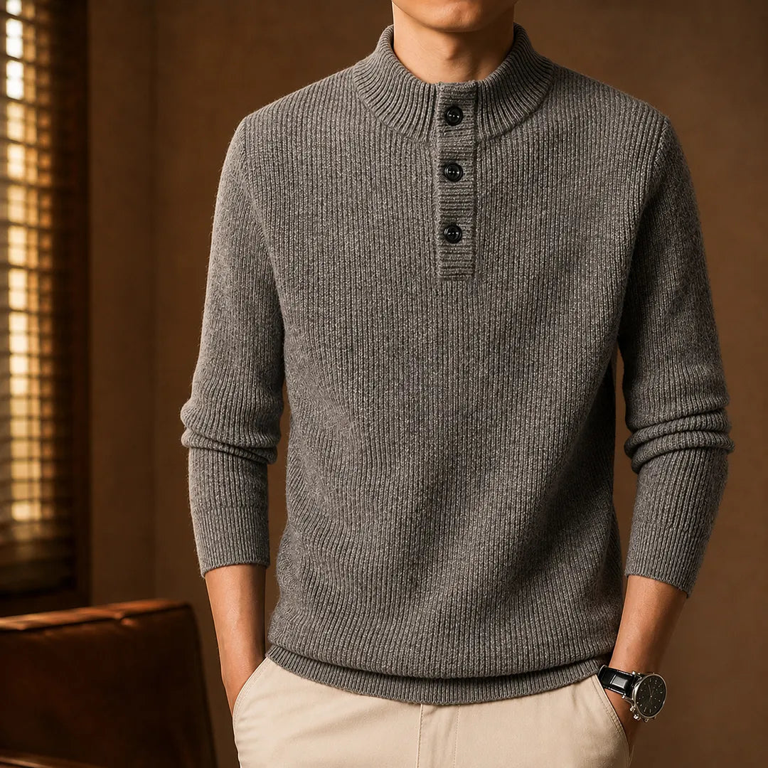 A man wearing a gray ribbed knit mock-neck sweater with three black buttons, paired with beige trousers, in a warm-toned indoor setting.